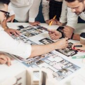 Four people review and discuss architectural floor plans and interior design photos laid out on a table, pointing and writing notes, with coffee cups and stationery nearby.