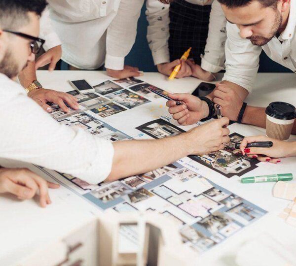Four people review and discuss architectural floor plans and interior design photos laid out on a table, pointing and writing notes, with coffee cups and stationery nearby.