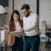 Two coworkers stand together in an office, smiling while looking at a tablet. The woman wears a brown blazer, and the man wears glasses and a white shirt. Modern office decor and plants are visible in the background.