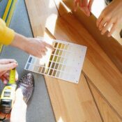 Three people compare paint color swatches to light wood flooring, one holding a tape measure and another pointing to the samples. Sunlight shines across the scene.