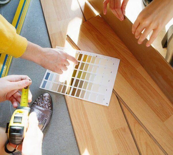 Three people compare paint color swatches to light wood flooring, one holding a tape measure and another pointing to the samples. Sunlight shines across the scene.