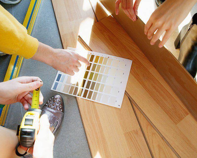 Three people compare paint color swatches to light wood flooring, one holding a tape measure and another pointing to the samples. Sunlight shines across the scene.