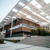 Modern building with wooden and glass exterior, large overhanging white louvered roof, stone steps leading up, and blossoming trees and greenery in the foreground. The sky is clear and bright.