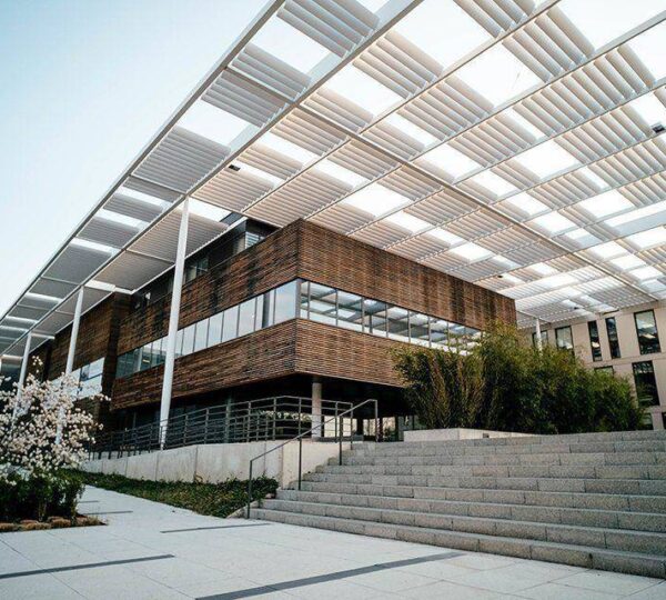 Modern building with wooden and glass exterior, large overhanging white louvered roof, stone steps leading up, and blossoming trees and greenery in the foreground. The sky is clear and bright.