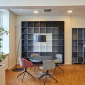 Modern office space with a round black table, four colorful chairs, a tall black floor lamp, empty shelving units, a wood floor, and a potted plant near a window. The room is well-lit and neatly organized.