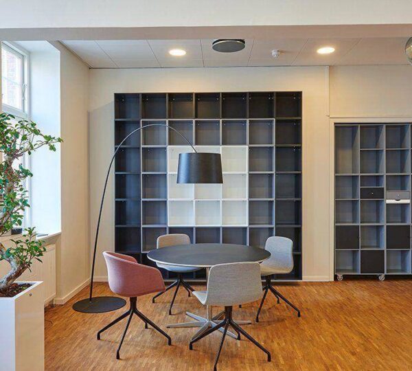 Modern office space with a round black table, four colorful chairs, a tall black floor lamp, empty shelving units, a wood floor, and a potted plant near a window. The room is well-lit and neatly organized.